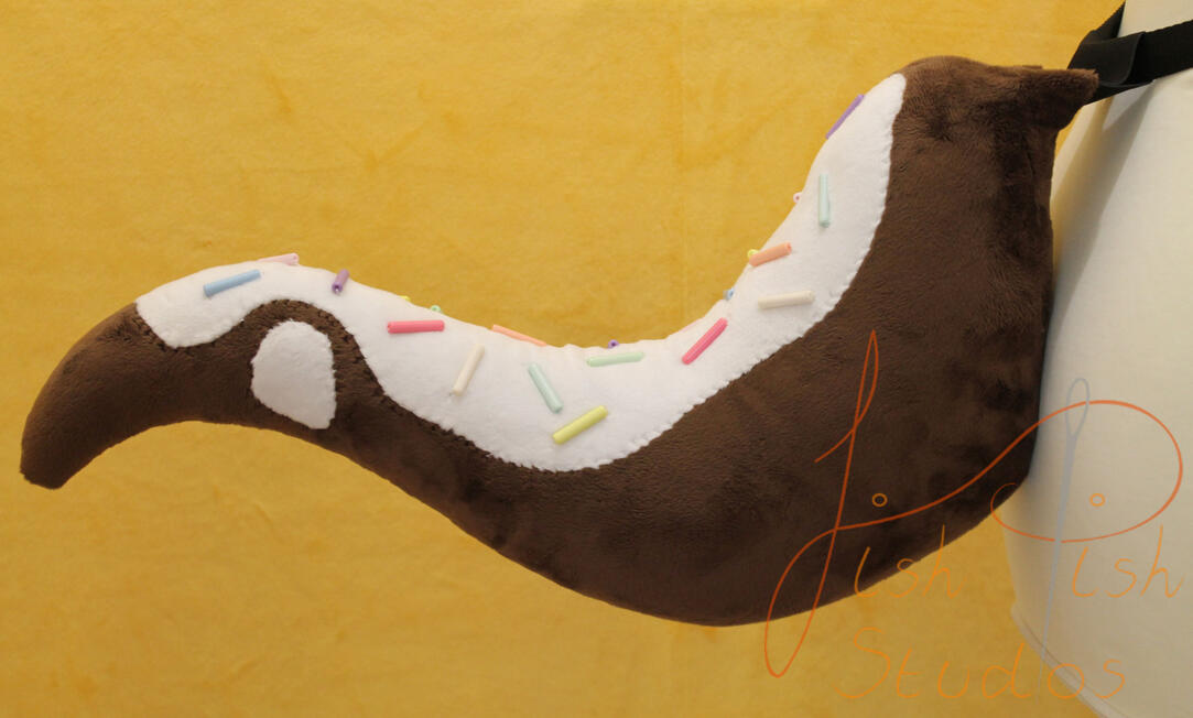 A brown coloured cake like raptor tail that sticks out. It has white icing and plastic bead sprinkle decorations. While it looks yummy, this cake cannot be eaten. It is made from minky for fursuiting, larp and therian costumes