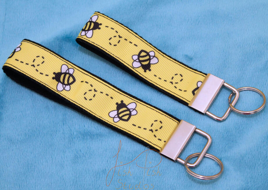 a cute photo showing two bee themed keyrings made of fabric. one is slightly bigger than the other. Both have little cartoon bees on them with a yellow background. handmade in the UK by fishpish studios